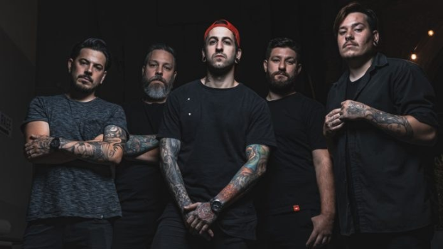 Eine Gruppe von Männern, die in einem dunklen Raum mit rechtsseitiger Beleuchtung stehen und wie bei einem Interview für Avenged Sevenfold aussehen.