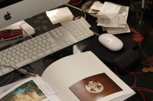 Ein Schreibtisch mit Monitor, Tastatur, Buch, Papieren, Maus, Münzen und einem Spielzeugauto.