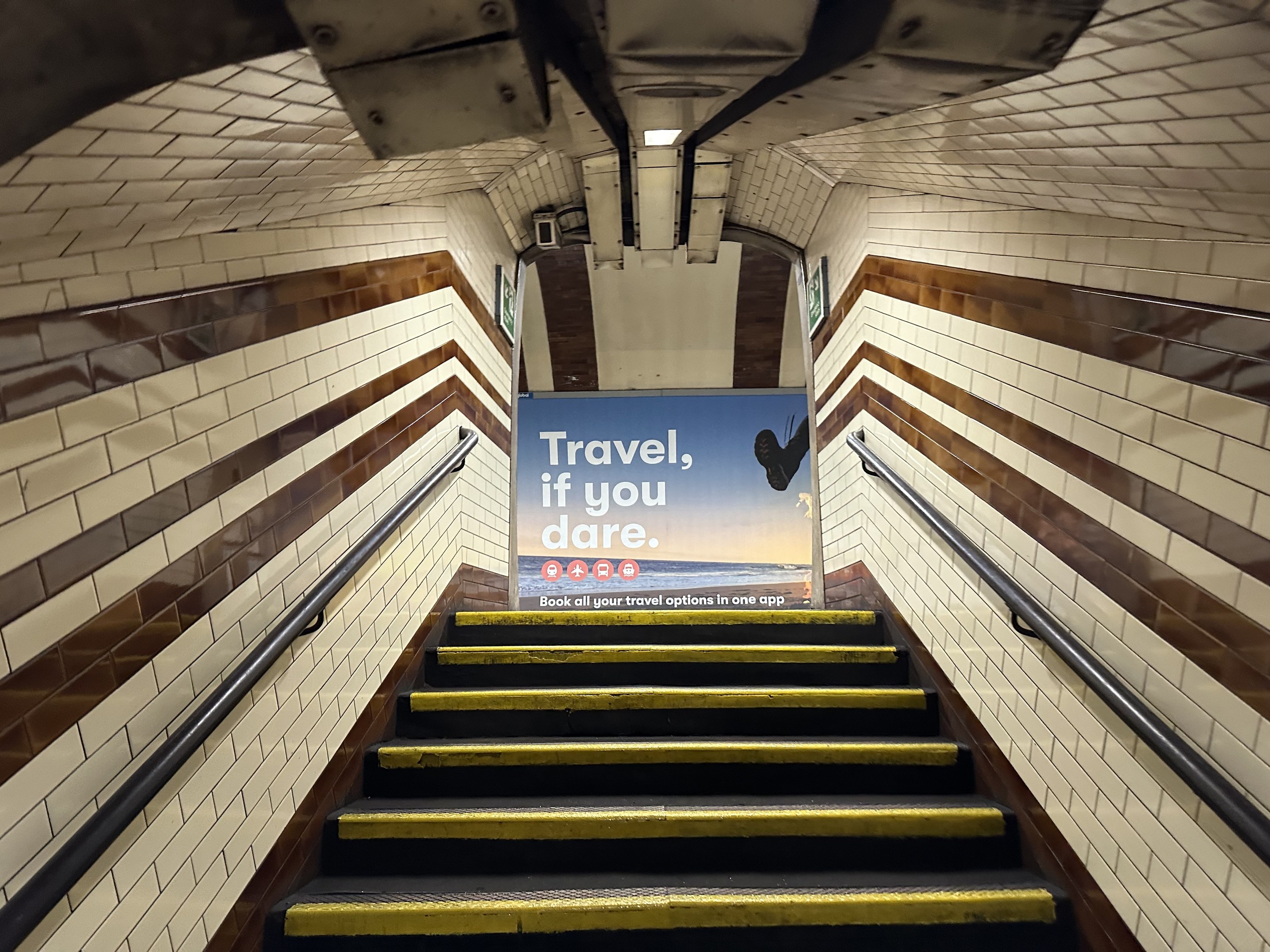 Eine Treppe in einer U-Bahn-Station mit einem 'Reise, wenn du dich traust'-Schild oben, flankiert von Geländern und sichtbaren Stationswänden.