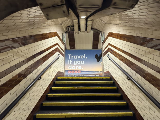 Eine Treppe in einer U-Bahn-Station mit einem 'Reise, wenn du dich traust'-Schild oben, flankiert von Geländern und sichtbaren Stationswänden.