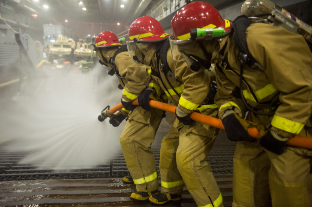 Feuerwehrleute in Helmen und Handschuhen halten Schläuche und spritzen Wasser auf ein Feuerwehrauto, mit Lampen, Geländern und Tafeln mit Text im Hintergrund.