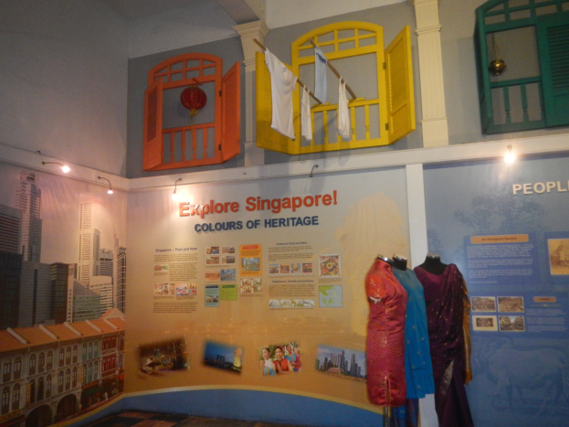 Ein Raum mit Schaufensterpuppen in Kleidern, eine Wand mit Text und Bildern, darunter "Erkunden Sie die Farben Singapurs der Tradition", Deckenlichter und ein Fenster mit Kleidung daran.