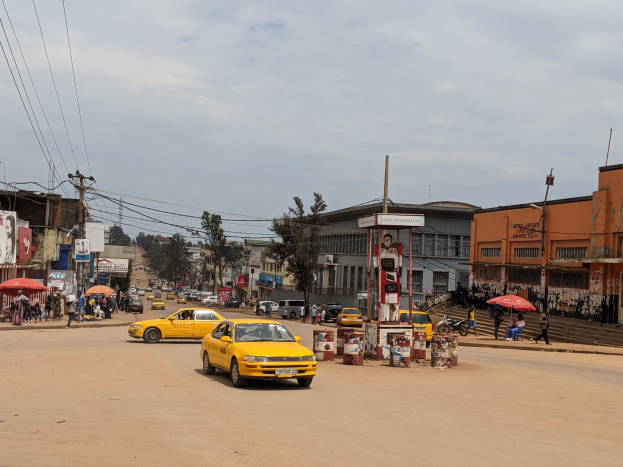 Ein gelbes Taxi fährt auf einer belebten Stadtstraße mit Gebäuden an den Seiten, während Fußgänger mit Schirmen auf dem Gehweg darunter gehen.