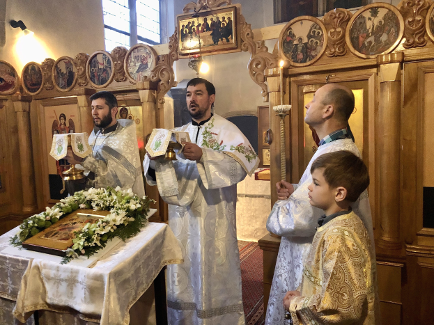Eine Gruppe von Menschen steht um einen Tisch in einer Kirche, hält Bücher in den Händen, mit einem Tuch und einem Blumenstrauß auf dem Tisch, und Fotorahmen, Lampen, ein Fenster und ein hölzernes Objekt im Hintergrund.