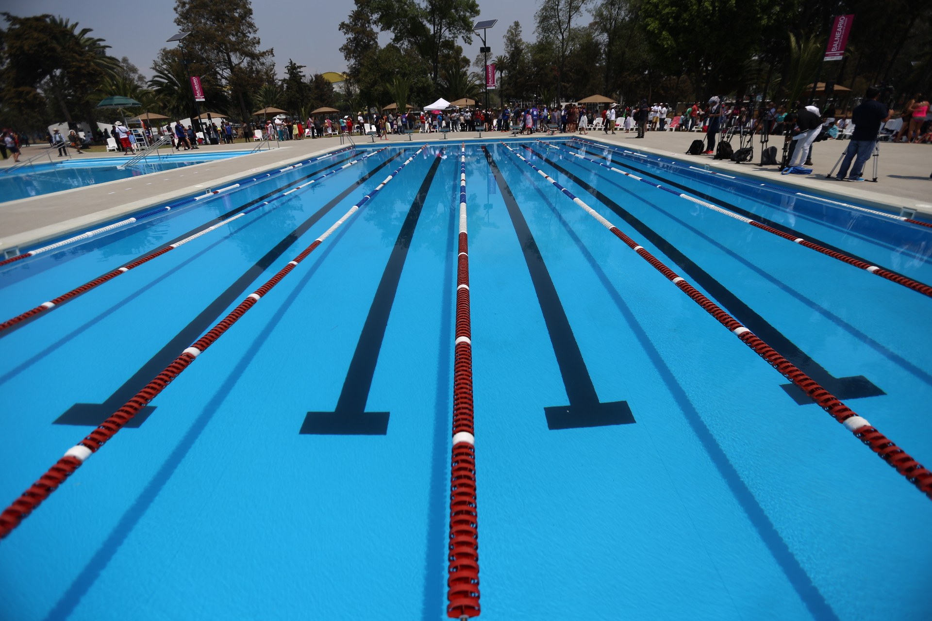 Großer Schwimmbad mit Bahnentrennungen, umgeben von Menschen mit Taschen und Gegenständen, mit Zelten, Transparenten, Pfosten, einem Schild und einem bewölkten Himmel im Hintergrund.