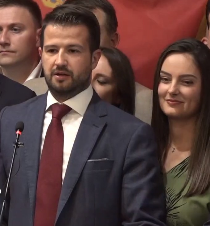 Ein Mann im Anzug spricht in ein Mikrofon an einem Rednerpult, mit einem Banner dahinter und einer aufmerksamen Menge davor.