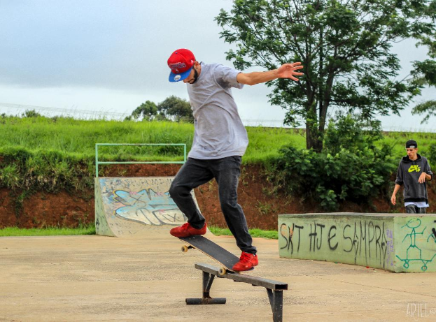 Eine Person fährt in der Mitte des Bildes Skateboard, mit Pflanzen, Bäumen, Gras und einer Böschung im Hintergrund, eine weitere Person steht rechts, ein Weg unten und der Himmel oben.