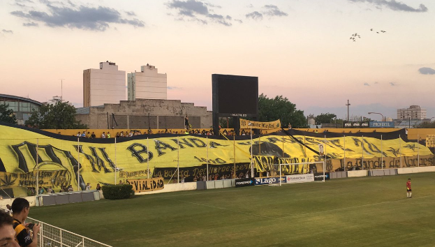 Ein Fußballfeld mit einer Gruppe von Menschen darauf, umgeben von einem Metallzaun, einem Torpfosten, einer Tafel mit der Aufschrift "Boca Juniors vs Real Sociedad", einigen Pflanzen, einem Bildschirm, einigen Pfählen, der Rinde eines Baums, einigen Bäumen, einigen Gebäuden und einem bewölkten Himmel.