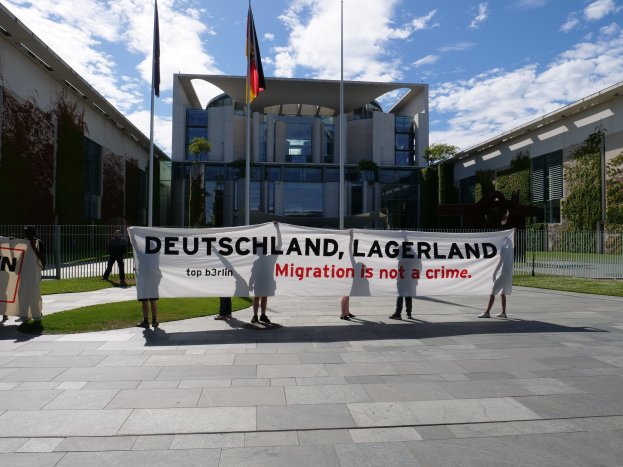 Gruppe von Menschen, die eine Tafel mit der Aufschrift "Deutschland, Lagerland Migration ist kein Verbrechen" vor einem Gebäude mit Fenstern halten, umgeben von Gras, einem Metallzaun, Pflanzen, Bäumen und Fahnenmästen.