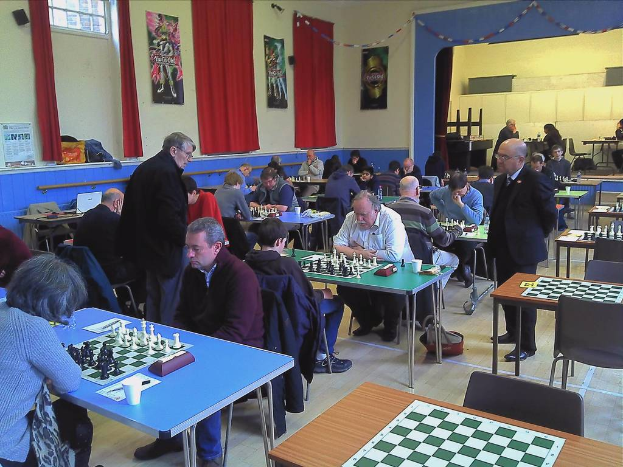 Zwei Personen spielen Schach an einem Tisch mit MÃ¼nzen auf dem Brett in einem groÃen Saal mit VorhÃ¤ngen, Plakaten, einem Fenster und einem Kalender an der Wand.