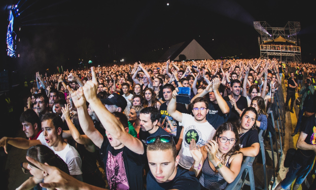 Eine große Menschenmenge auf einem Musikfestival mit erhobenen Händen, einige sitzen und andere stehen, mit einem Metallzaun auf der rechten Seite, Gebäuden, Lichtern und einem dunklen Himmel im Hintergrund und einem Mann, einer Frau und anderen Menschen im Vordergrund.