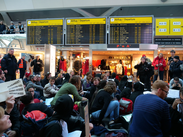 Eine große Gruppe von Menschen in einem Flughafen, einige sitzen mit Taschen und Papieren, andere stehen, mit Texttafeln und Schaufensterpuppen im Hintergrund, was auf eine Protestaktion hinweist.