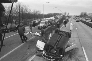 Schwarz-weißes Bild eines umgestürzten Autos am Straßenrand mit beschädigten Fahrzeugen in der Nähe und Menschen, die sich darum versammeln, vor einem Hintergrund aus Laternenpfählen, Bäumen, einer Brücke und Himmel.