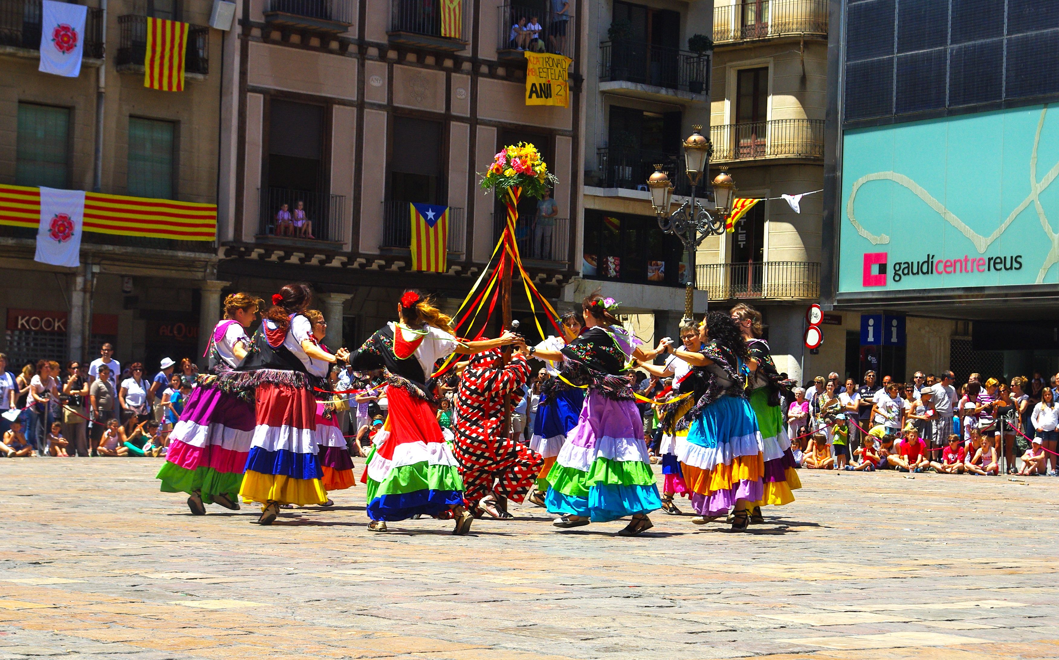 Eine Gruppe von Frauen in bunten Kleidern tanzt auf der Straße, umgeben von einer Menge Menschen, mit Gebäuden, Geländern, Balkonen, Flaggen, einem Laternenmast und einer Tafel mit Aufschrift im Hintergrund.