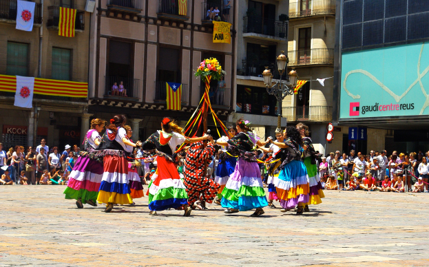 Eine Gruppe von Frauen in bunten Kleidern tanzt auf der Straße, umgeben von einer Menge Menschen, mit Gebäuden, Geländern, Balkonen, Flaggen, einem Laternenmast und einer Tafel mit Aufschrift im Hintergrund.