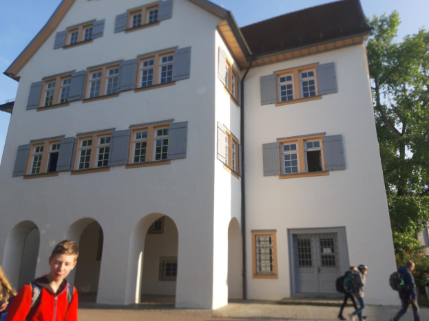 Eine Gruppe von Menschen, einige mit Taschen, geht vor einem großen weißen Gebäude mit Fenstern und Türen vorbei, umgeben von Bäumen unter einem klaren blauen Himmel, was darauf hindeutet, dass sie auf dem Weg zu einer Schule in Deutschland sind.