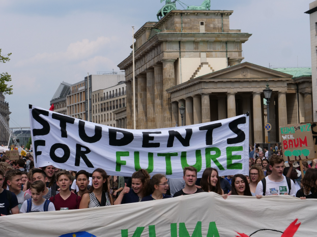 Schüler marschieren in Berlin mit einem bunt bemalten 'Students for Future'-Schild vor einer Kulisse aus Gebäuden, Bäumen und Himmel.