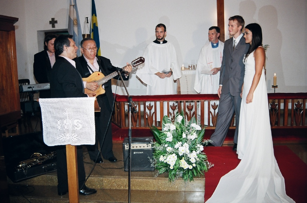 Eine Hochzeitsszene in einem Raum mit zwei Pastoren, einem Brautpaar und drei Musikern; ein Podium mit Stoff, ein Tisch mit Kerzen hinter den Pastoren, ein Kerzenhalter hinter dem Paar und verschiedene Gegenstände auf dem Boden wie ein Bukett, Lautsprecher und Taschen; Flaggen, eine Wand mit einem Kreuz, ein Schrank und ein Stuhl im Hintergrund.