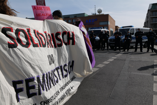 Eine Gruppe von Menschen, die mit einem "Solidarität und Feminismus"-Schild die Straße entlangmarschieren, mit parkenden Fahrzeugen und Gebäuden im Hintergrund unter einem klaren blauen Himmel.