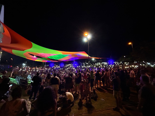 Eine große Menschenmenge versammelt sich nachts vor einem Zelt, das von bunten Lichtern und Laternen beleuchtet wird, mit einem Gebäude auf der linken Seite und dem Nachthimmel im Hintergrund, der die Atmosphäre des 2018er Kunstfestivals einfängt.