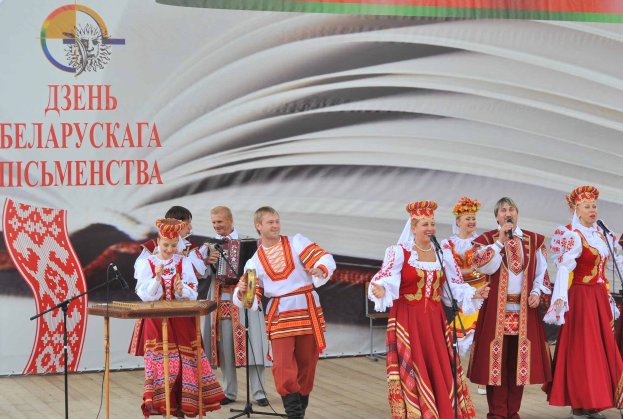 Eine Gruppe von Menschen in rot-weißer Kleidung performs auf der Bühne mit Mikrofonen und Instrumenten, mit einem 'Ukrainischer Folklore-Festival'-Banner im Hintergrund.