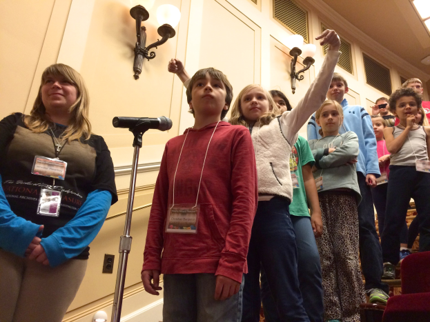 Gruppe von Kindern singt an einem Mikrofon mit einer Frau neben ihnen, Stühle auf der rechten Seite und beleuchtete Wand im Hintergrund.