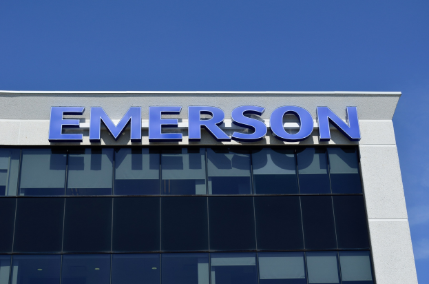 Emerson-Logo an einer Gebäudefassade mit dem Text "Emerson investiert 1,5 Milliarden US-Dollar" vor einem klaren blauen Himmel.