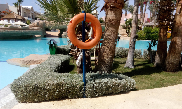 Schwimmbad mit einem Rettungsring in der Mitte, umgeben von grünem Gras, Pflanzen und Bäumen, mit Gebäuden, Sonnenschirmen und einem klaren blauen Himmel im Hintergrund.