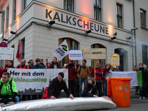 Eine Gruppe von Menschen mit Schildern und Plakaten steht vor einem Gebäude, mit zwei Personen im Vordergrund und einem Müllimerker rechts, während einer Demonstration in Deutschland.