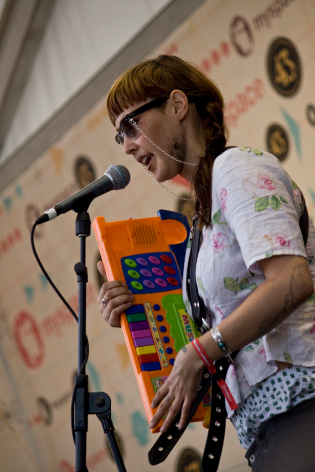 Eine Frau in einem weißen Hemd und Sonnenbrille singt in ein Mikrofon auf einem Ständer und hält dabei eine Piano, mit einem Plakat an der Wand dahinter.