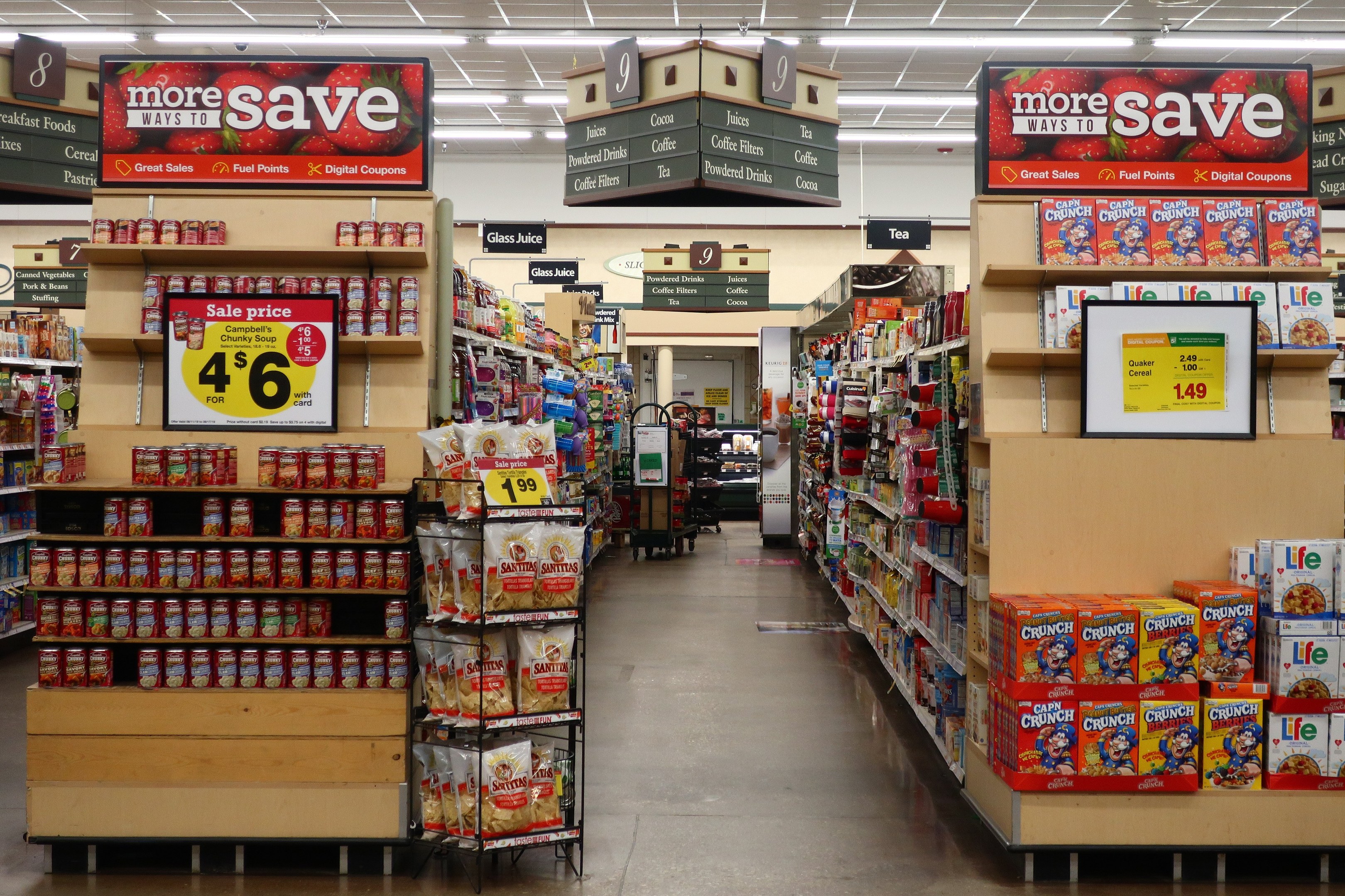 Ein Innenraum eines Lebensmittelladens mit verschiedenen Lebensmitteln auf Regalen, Regalen und dem Boden, mit Deckenleuchten und Informationsschildern, mit einer Person im Hintergrund.