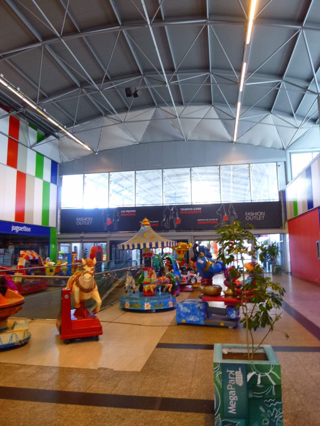 Großer Indoor-Spielplatz in einem Einkaufszentrum mit verschiedenen Fahrgeschäften, einem Topf mit einer Pflanze, verstreuten Spielzeugen, Informationsschildern, Deckenleuchten und Metallstützen.