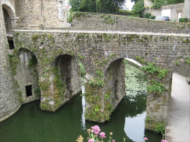 Eine Brücke spannt sich über das Bild mit einer Straße, einem Baum und Wasser darunter, einem Auto und einer Wand in der rechten oberen Ecke und einem Gebäude auf der linken Seite.