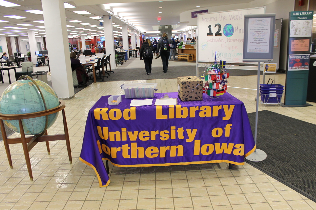 Eine Ausstellung mit einem Tisch, auf dem verschiedene Landesflaggen, eine Box, Papiere und ein Banner ausgebreitet sind, eine Kugel links daneben, ein Anschlagbrett rechts daneben mit ein paar Menschen in der Nähe und viele Tische und Stühle im Hintergrund.