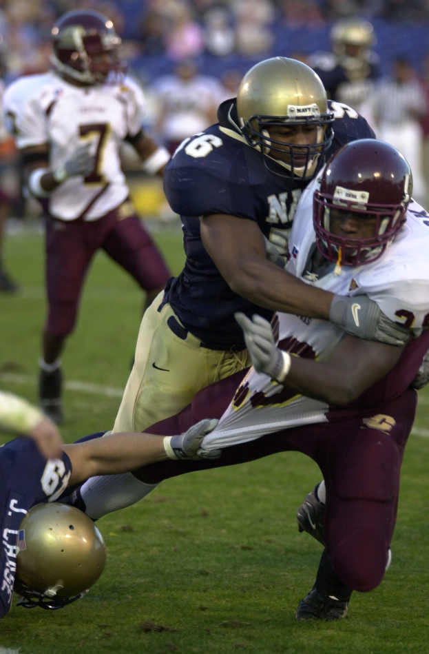Zwei Fußballspieler in Helmen und Uniformen kämpfen auf dem Feld, während andere zuschauen.