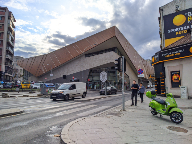 Ein grüner E-Scooter steht auf dem Seitenstreifen neben einem Gebäude, mit Fahrzeugen auf der Straße, Fußgängern auf dem Gehweg und Ampelmasten, während Gebäude mit Fenstern, Bäume und ein bewölkter Himmel im Hintergrund zu sehen sind.