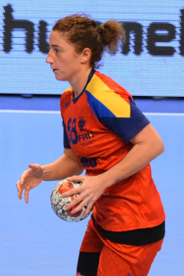 Eine Frau in einem orange-blauen Trikot hält einen Fußball, im Hintergrund ist ein Bildschirm mit Text zu sehen.