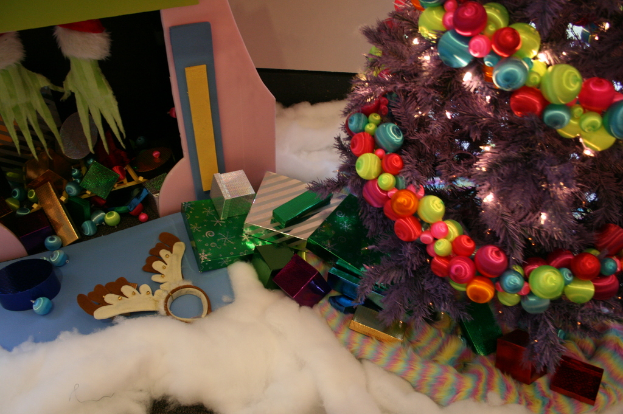 Drinnen steht rechts ein geschm├Ęckter Weihnachtsbaum mit Lichterkette und Schmuck, umgeben von Geschenkboxen und Deko-Artikeln, mit einer H├╝tte und Watte oben links und Wolle unten.
