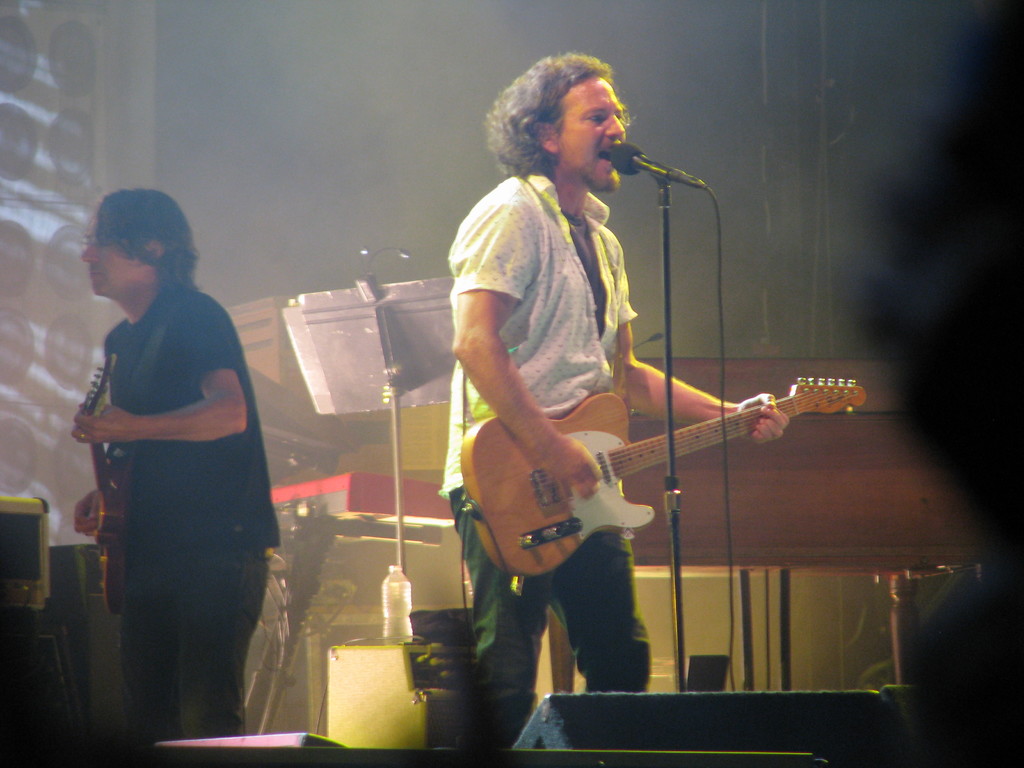 Ein Mann in einem weißen Hemd spielt Gitarre und singt in ein Mikrofon auf der Bühne, während ein anderer Mann in einem schwarzen Hemd neben ihm Gitarre spielt; ein Piano, Kabel, Lautsprecher und Lichter sind im Hintergrund zu sehen.