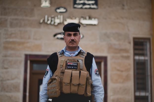 Polizist in Uniform und Mütze vor einem Gebäude mit Türen, Fenstern und Text an der Wand.