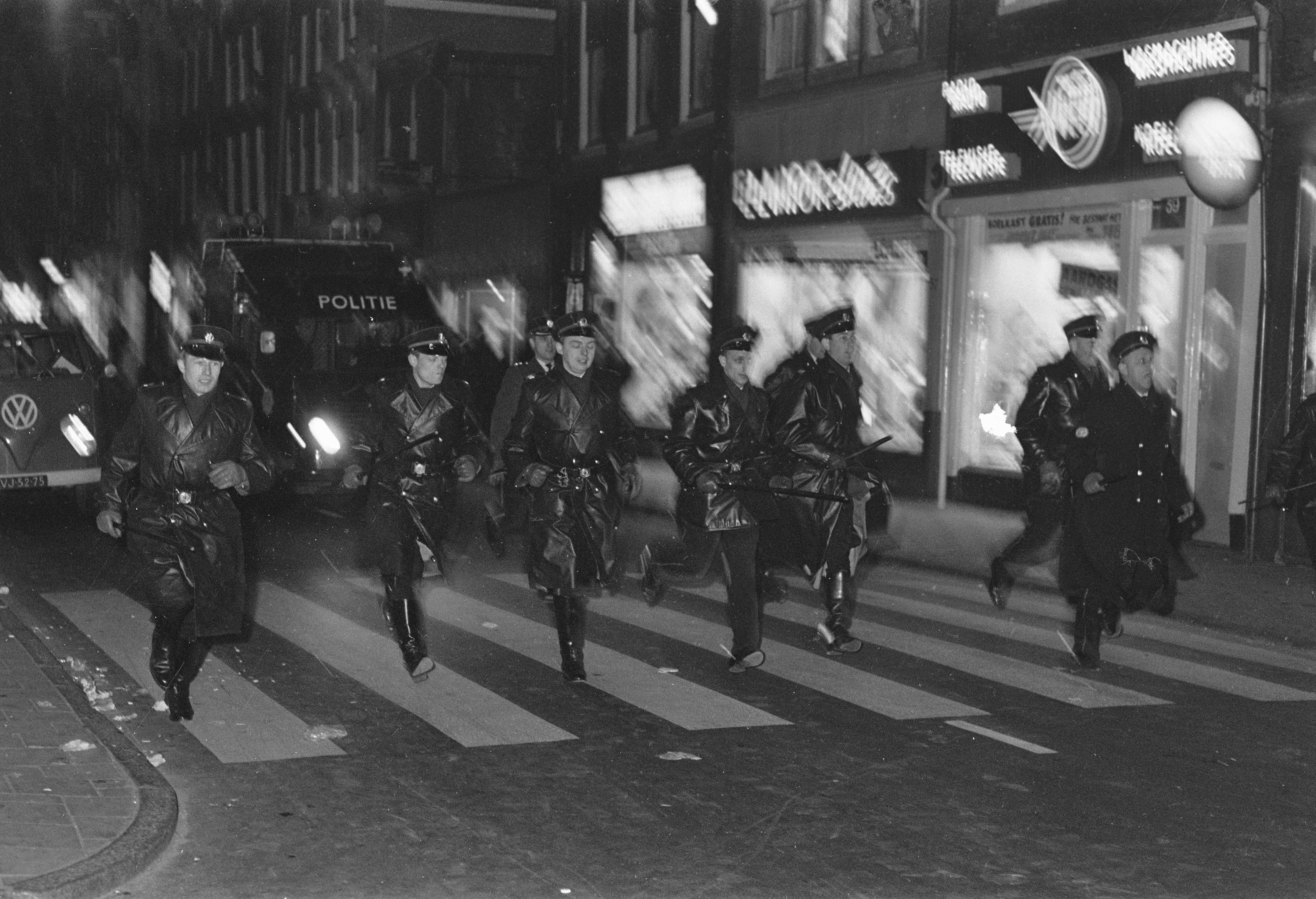 Schwarzes und weißes Bild von Polizisten in Uniform, die nachts über eine Straße gehen, einige tragen Waffen, mit Fahrzeugen und Gebäuden im Hintergrund.