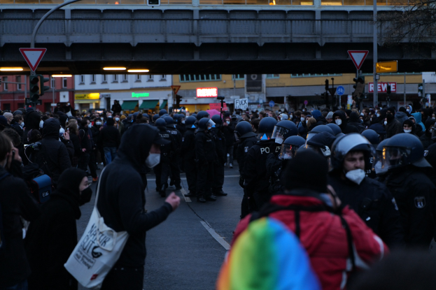 Eine große Gruppe von Menschen vor einer Reihe von Polizisten, einige mit Helmen und Taschen oder Kameras, mit Gebäuden, Schildern, Laternen, Verkehrszeichen, einer Brücke und einem Baum im Hintergrund.