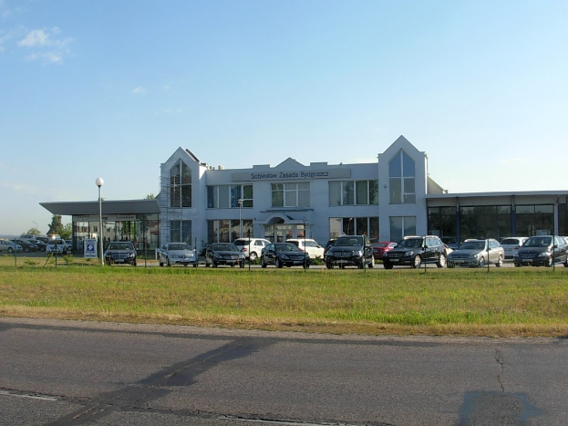 Ein Autohaus mit parkenden Autos vorne, umgeben von Gebäuden, Straßeninfrastruktur, fahrenden Fahrzeugen, Bäumen und einem bewölkten Himmel.