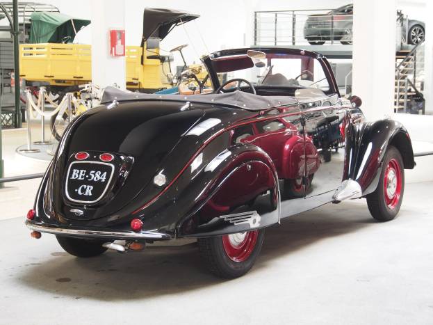 Schwarzes und rotes Auto in einer Garage mit anderen Fahrzeugen, Pfählen, Ketten und Gegenständen geparkt, mit einem Feuerlöscher an der Wand im Hintergrund.