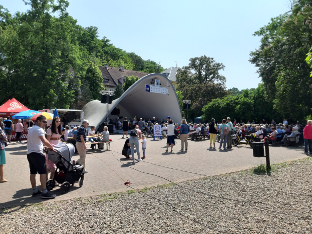 Menschenmenge bei Festival-Eröffnung, einige sitzen auf Bänken, andere stehen, mit einem Kinderwagen im Vordergrund, Zelte links, Mülltonne rechts und Gebäude, Bäume und klarer blauer Himmel im Hintergrund.
