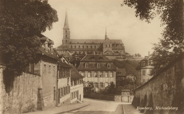 Schwarz-weißes Foto einer Stadtstraße in Bamberg, Deutschland, mit einer Kirche im Hintergrund, flankiert von Mauern und gesäumt von Bäumen.