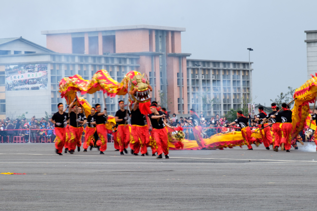 Eine Gruppe von Menschen in roten und gelben Kostümen führt einen chinesischen Neujahrsumzug auf einer Straße auf, während Zuschauer, Gebäude, Bäume, Laternenmasten und ein Banner mit Text und Bildern im Hintergrund zu sehen sind, unter einem sichtbaren Himmel.
