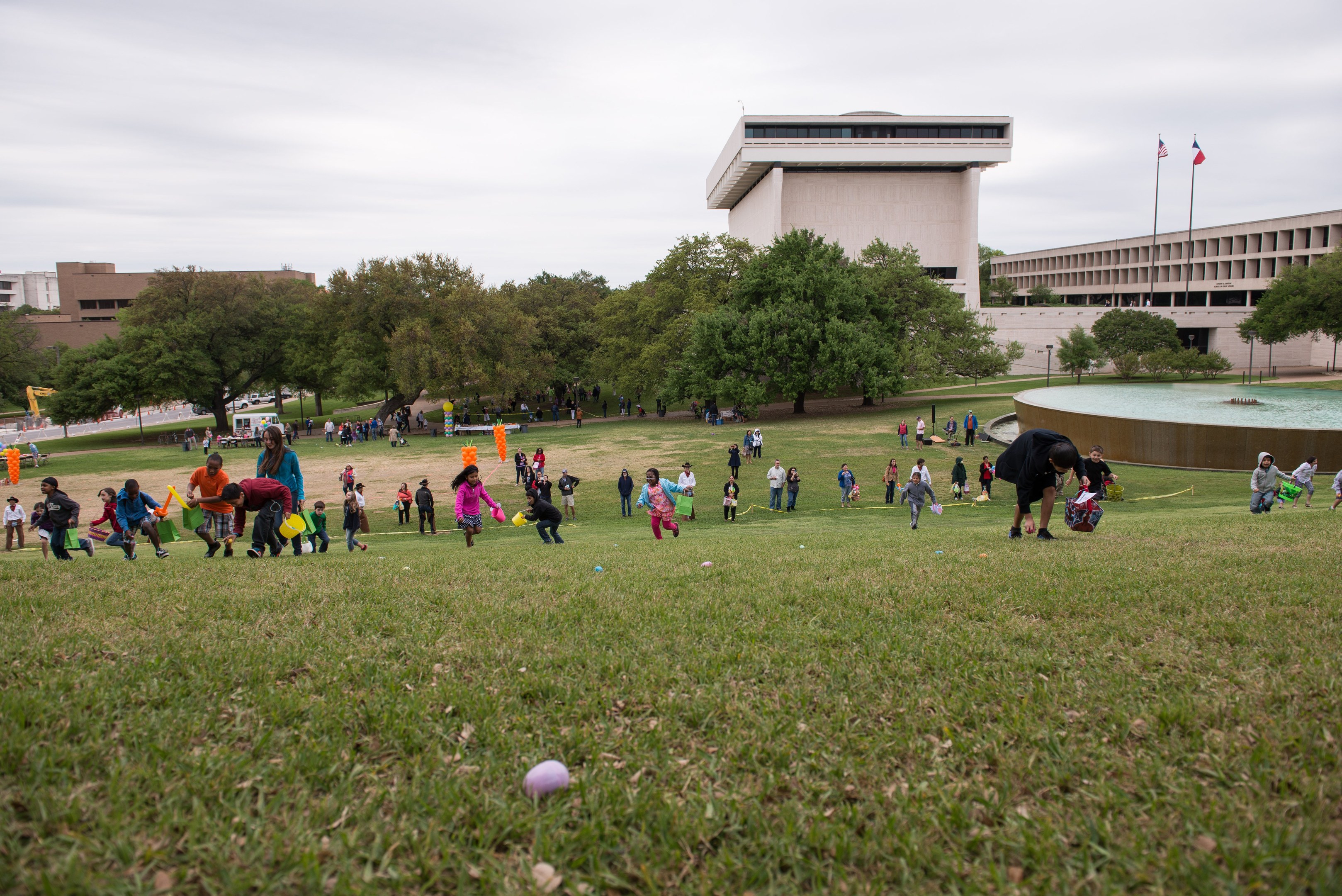 Gruppe von Menschen, die mit Ostereiern auf einer grünen Wiese mit Bäumen, Gebäuden, Fahnen, einem Brunnen und einem bewölkten Himmel spielt.