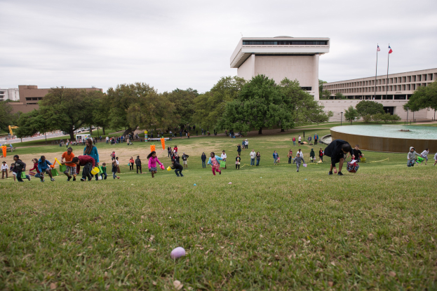 Gruppe von Menschen, die mit Ostereiern auf einer grünen Wiese mit Bäumen, Gebäuden, Fahnen, einem Brunnen und einem bewölkten Himmel spielt.
