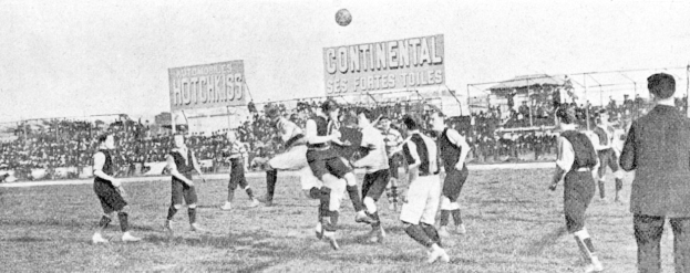 Ein Schwarz-Weiß-Bild einer Gruppe von Männern, die auf einem Feld Fußball spielen, mit einem Ball in der Luft und Zuschauern im Hintergrund, mit Tafeln, die wahrscheinlich textbezogen zum Spiel sind.